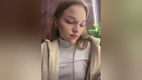 Media: Video of a young woman with light skin and straight, shoulder-length blonde hair, wearing a white turtleneck and beige cardigan, looking contemplative, indoors with a green potted plant and wooden wall behind.