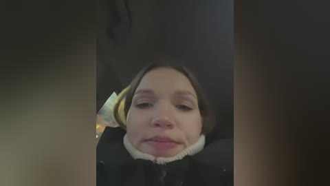 Media: A video of a young woman with light skin and brown hair, wearing a black jacket, seated in a dimly lit car, with a blurred background.