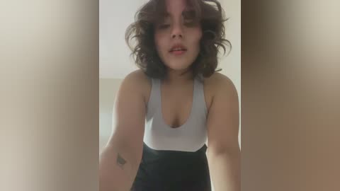 Media: Video of a young woman with curly black hair, wearing a grey tank top with a deep neckline, and black high-waisted pants, standing in a beige doorway.