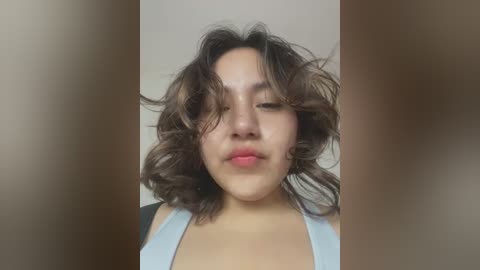 Media: Video of a young Asian woman with shoulder-length, wavy brown hair, wearing a light blue tank top. She has a neutral expression and stands against a plain, beige background.