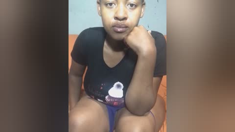 Media: Video of a young Black woman with medium-brown skin, sitting on a couch, wearing a black T-shirt with a cartoon character, and blue panties. She has short hair and a neutral expression.