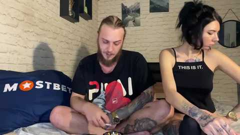 Media: Video of a bearded, tattooed man and a woman with black hair and tattoos sitting on a bed, both in black T-shirts, in a cozy, dimly lit room with exposed brick walls.