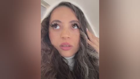 Video of a young woman with long, wavy brown hair, wearing a white turtleneck sweater. She has a light brown complexion, expressive brown eyes, and pink lips. She is looking up with a contemplative expression, standing in a room with beige walls and a wooden door.