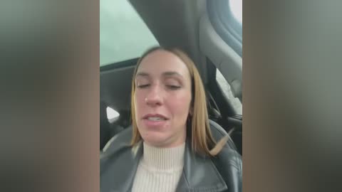 Media: A video of a Caucasian woman with light skin, long blonde hair, wearing a black leather jacket over a cream sweater, seated in a car with her eyes closed, seemingly asleep.