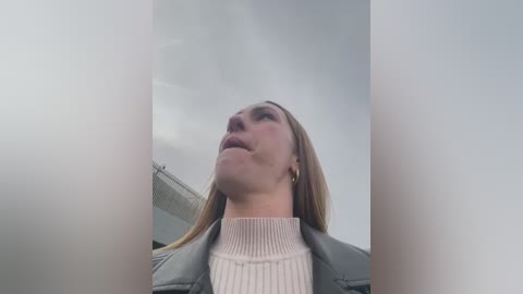 Media: A video of a blonde woman with light skin and a slightly pensive expression, wearing a light-colored sweater and gray jacket, taken from a low angle, with a cloudy sky background.