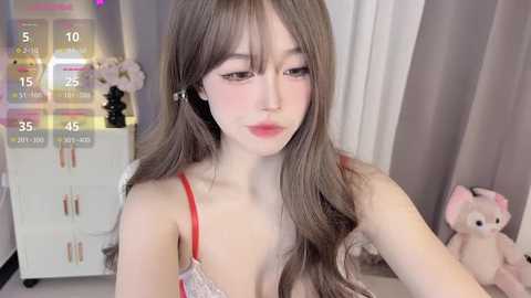 Media: Video of a young woman with long brown hair, fair skin, and red lipstick, wearing a white lace bra, in a cozy room with a white dresser, pink teddy bear, and soft lighting.