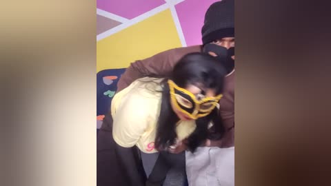 Media: Video of a young woman with long black hair, wearing a yellow mask, held by a man in a brown jacket and black hat in a colorful, geometrically patterned room.