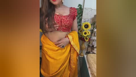 Media: Video of a young woman with light skin and long brown hair, wearing a red lace top and a vibrant yellow sari, standing indoors beside sunflowers and a tiled wall.