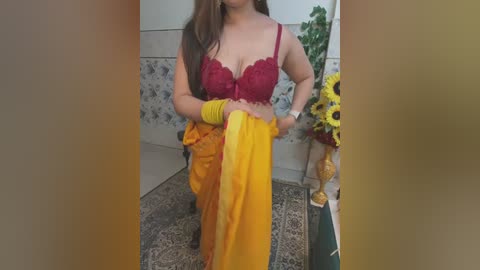 Media: A video of a fair-skinned woman in a red lace bra, holding a yellow sari, with floral wallpaper and a sunflower vase in the background.