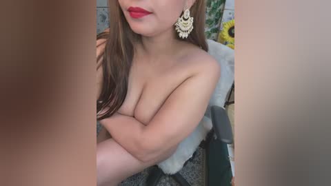 Media: Video of a topless, fair-skinned woman with long brown hair, wearing large gold earrings and red lipstick, sitting on a chair with her arms crossed, partially obscured by two large, brown hands. Background includes a sunflower and tiled wall.