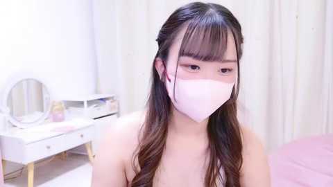 Media: Video of an Asian woman with long, straight black hair and a white mask, standing in a soft-pink bedroom with a vanity table, mirror, and a bed.