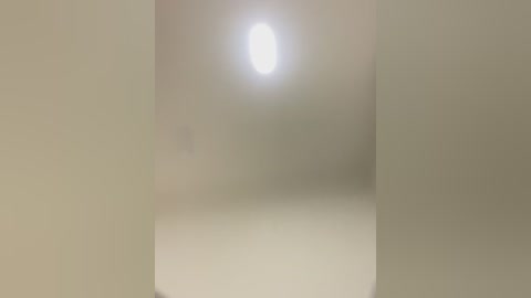 Media: A video of a minimalist, empty room with soft, diffused lighting from a central ceiling bulb, creating a serene, monochromatic ambiance. The walls and floor are smooth, light beige, emphasizing the calm and sparseness.