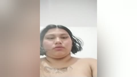 Media: Video of an East Asian woman with medium skin tone, shoulder-length black hair, and a tattoo on her chest, standing topless in a white bathroom.