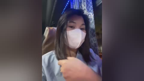 Media: A video of an Asian woman with long black hair, wearing a white surgical mask, sitting in a dimly lit room with blue LED lights. She is holding a phone, possibly taking a selfie.