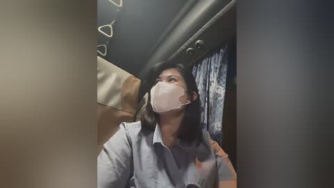 Media: Video of a young woman in a light blue shirt, wearing a white face mask, seated on a plane. The overhead compartment is visible, with a beige garment hanging inside.