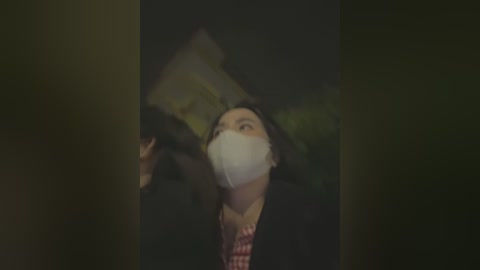 Media: A dimly lit video of a woman with a white mask and dark hair, wearing a red and black checkered shirt, looking upward in a dark, possibly urban, setting.