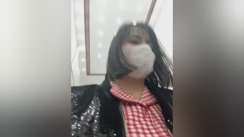 Media: A video shows a person with short black hair, wearing a white face mask, a red plaid shirt, and a black sequined jacket, standing indoors with a blurred background.