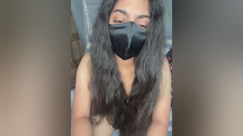 Media: Video of a topless woman with long, wavy dark hair, wearing a black face mask, seated indoors, with a blurred background.