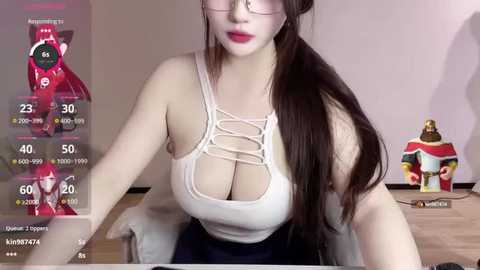 Media: Video of an Asian woman with long dark hair, wearing a revealing white top with lace details, sitting at a desk. Background shows a computer screen displaying stats, and a plush toy and other objects.