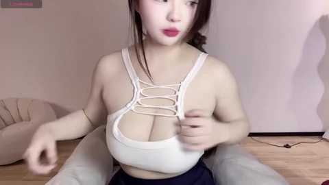 Media: A video of a light-skinned woman with long, dark hair, wearing a revealing white lace top that accentuates her large breasts and a navy skirt, sitting on a beige chair in a minimalist room with wooden floors.