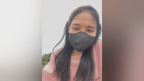 Media: Video of a young Asian woman with long black hair, wearing a black face mask and a pink sweatshirt, standing against a plain white wall, with a blurred green plant in the background.