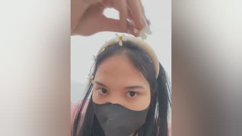 Media: Video of a young Asian woman with long black hair, wearing a black face mask and a light headband, being held by a hand, indoors with soft lighting.