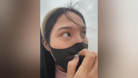 Media: Video of a young Asian woman with straight black hair, wearing a black mask, holding a hand to her mouth, in a blurred, beige-toned background.