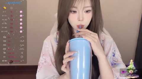 Media: Video of an East Asian woman with long brown hair, wearing a white kimono, drinking from a blue cup, with a calendar and a small toy in the background.