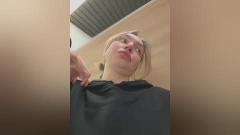 Media: Video of a young Caucasian woman with fair skin, light brown hair, and a black t-shirt, captured from a low angle, emphasizing her upturned mouth in a playful manner. Background includes beige walls and a ceiling with dark panels.