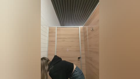 Media: Video of a narrow, beige-tiled restroom with a woman wearing a black hoodie and jeans, facing away, her long brown hair visible. The ceiling features a dark wood panel.