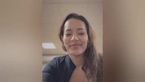 Video of a young woman with medium skin tone and long dark hair, wearing a black top, smiling slightly, in a neutral-toned room with a blurred background.