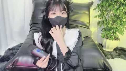 Media: A video of an Asian woman with long black hair and pale skin, wearing a black face mask and maid outfit, sitting on a black leather couch. She holds a remote control in her hand, with a potted plant and white curtains in the background.