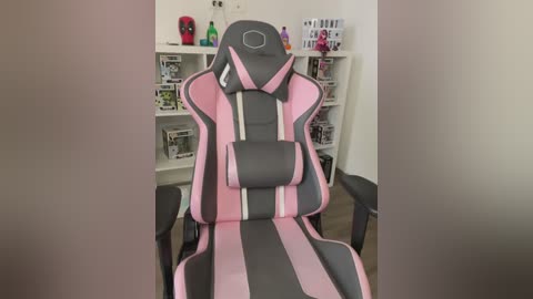 Media: Video of a pink and gray gaming chair with a padded headrest and lumbar support in a well-lit room, featuring a white bookshelf filled with gaming posters and decor.