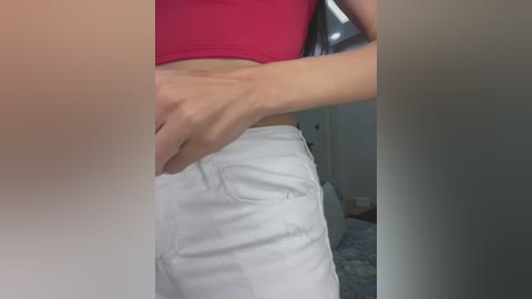 Media: Video of a woman's midsection, wearing a red crop top and white shorts, standing indoors. She adjusts her waistband, with her hand visible. Background shows a dimly lit room with a bed and a door.