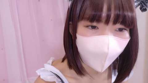 Media: A video of an Asian woman with straight, shoulder-length brown hair and bangs, wearing a white face mask, black lace headband, and a white off-shoulder dress. She has fair skin and is indoors against a pink background.