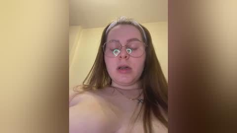 Media: A video of a young Caucasian woman with long brown hair, wearing glasses, a headband, and a necklace, standing topless against a beige wall, her expression neutral.