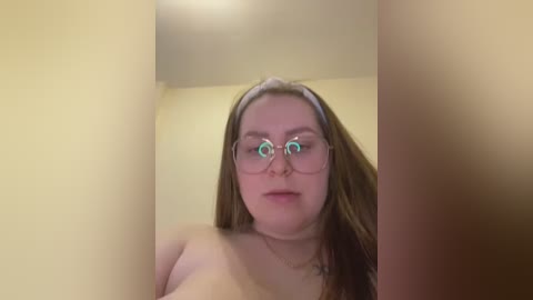 Media: Video of a fair-skinned woman with long brown hair, wearing round glasses, standing naked in a yellow-painted room. Her expression is neutral, and she appears to be in her late 20s.