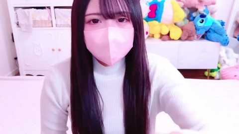 Media: A video of a young Asian woman with long black hair, wearing a light blue mask, white sweater, and white pants, sitting in a brightly lit, child-friendly room with stuffed toys and shelves.