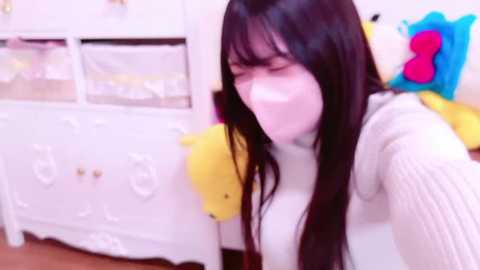 Media: Video of a young woman with long black hair, wearing a white sweater, and a white face mask, standing in a brightly lit room with a white dresser, colorful plush toys, and a soft carpet.