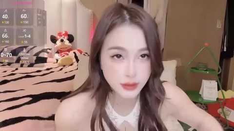 Media: Video of a young Asian woman with long brown hair, wearing a white top, sitting on a zebra-printed bed in a cozy room with a green toy cart, stuffed toys, and a wall clock.