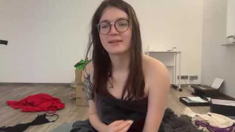 Media: Video of a young woman with glasses, long dark hair, and a black dress, sitting on a wooden floor in a cluttered room with scattered clothes and boxes.