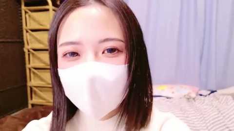 Media: Video of an East Asian woman with light skin, long black hair, and wearing a white face mask, standing indoors against a light blue curtain backdrop. She appears calm.