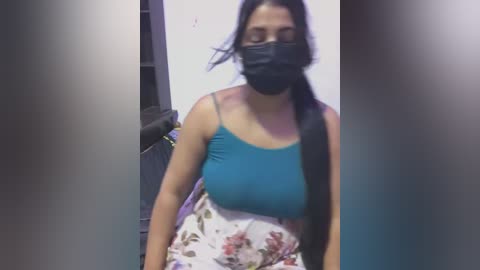 Media: A video shows a woman with long black hair, wearing a black face mask and a turquoise tank top with floral shorts. She appears to be walking through a dimly lit room, possibly an office or hallway.