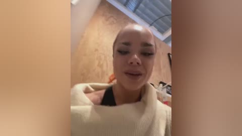 Media: Video of a bald, fair-skinned woman with black eyeliner, wearing a beige sweater, indoors with wooden walls and a blue ceiling fan.