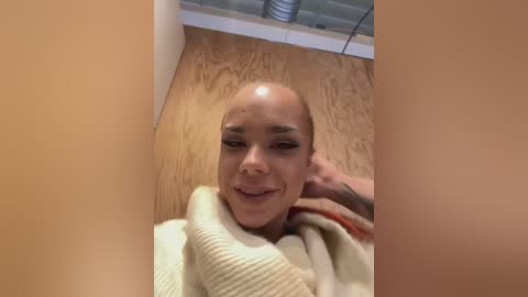 Media: Video of a bald, smiling woman with light skin, wearing a beige sweater, lying on a wooden floor with a white baseboard.