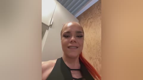 Media: Video of a smiling, bald, fair-skinned woman with long red hair, wearing a black halter top, standing in a narrow hallway with white and wooden walls.