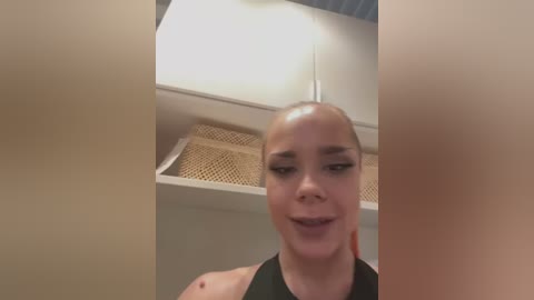 Media: Video of a smiling, light-skinned woman with a bald head, wearing a black top, standing in a modern, white kitchen with woven baskets on a shelf.