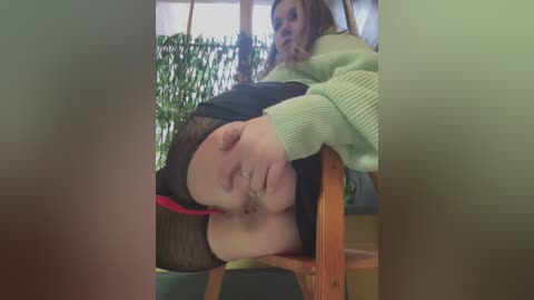 Media: Video of a woman with a bald head, wearing a green sweater and black sheer tights, lying on a wooden chair, with a blurry background featuring a potted plant and a window.