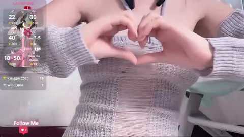 Media: Video of a fair-skinned woman in a knitted grey sweater, forming a heart shape with her hands. The background shows a bathroom with a white sink and a mirror.