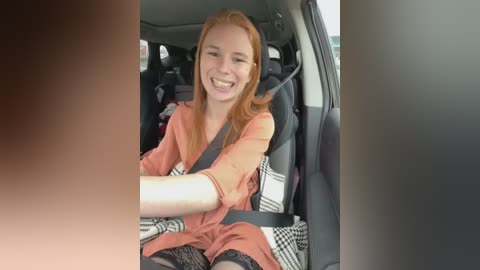 Media: Video of a smiling young woman with long, straight red hair, wearing a coral dress and black stockings, seated in a car with a gray seatbelt, black checkered-patterned seat cover, and a blurred background.